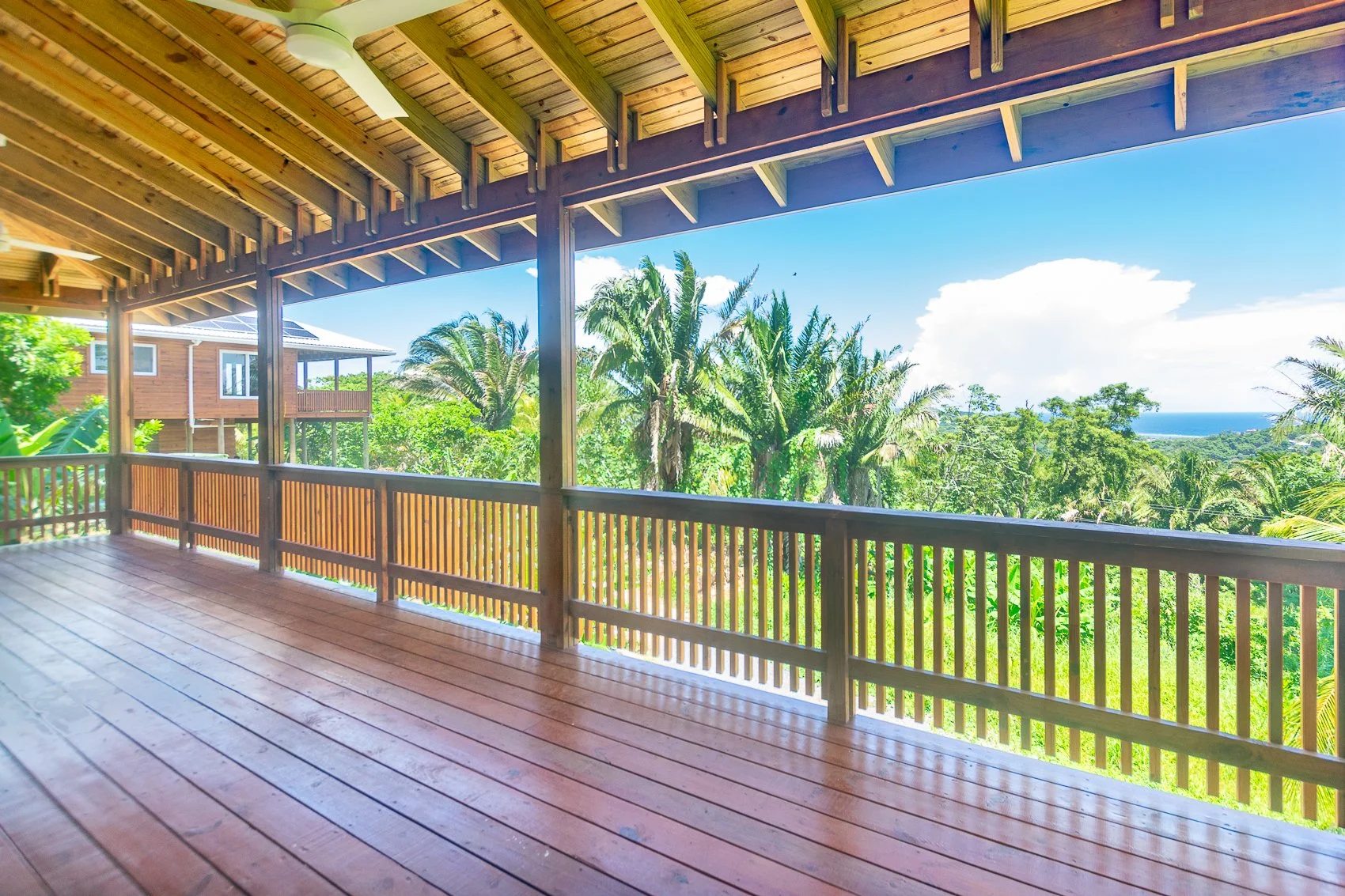 View from a wooden balcony overlooking lush tropical trees and a distant ocean under a blue sky with clouds.