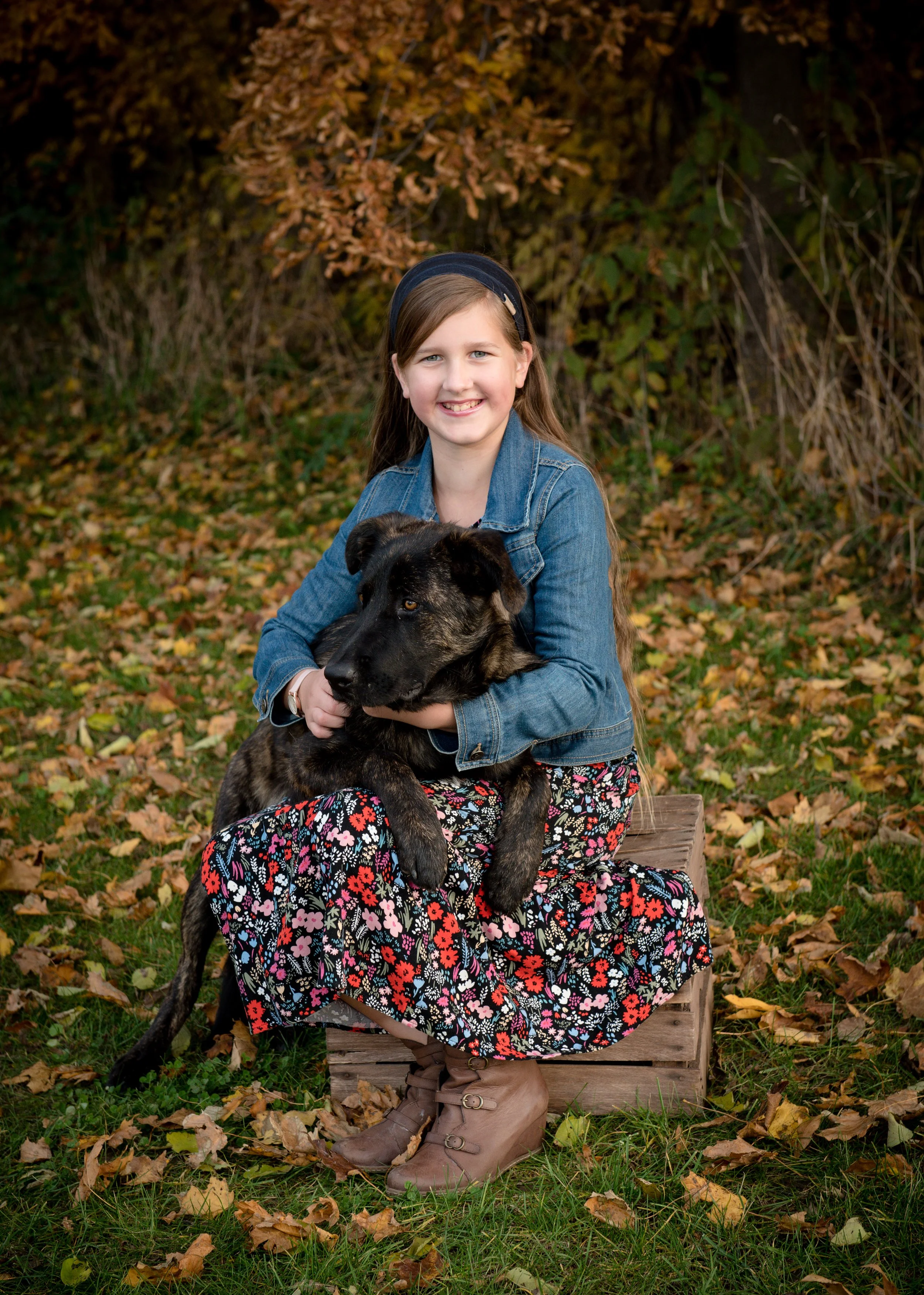 A young girl with light brown hair, wearing a denim jacket, floral skirt, brown boots, and a black headband, sitting on a wooden crate outdoors surrounded by fallen autumn leaves. She is holding a large, brindle-coated dog and smiling at the camera, 