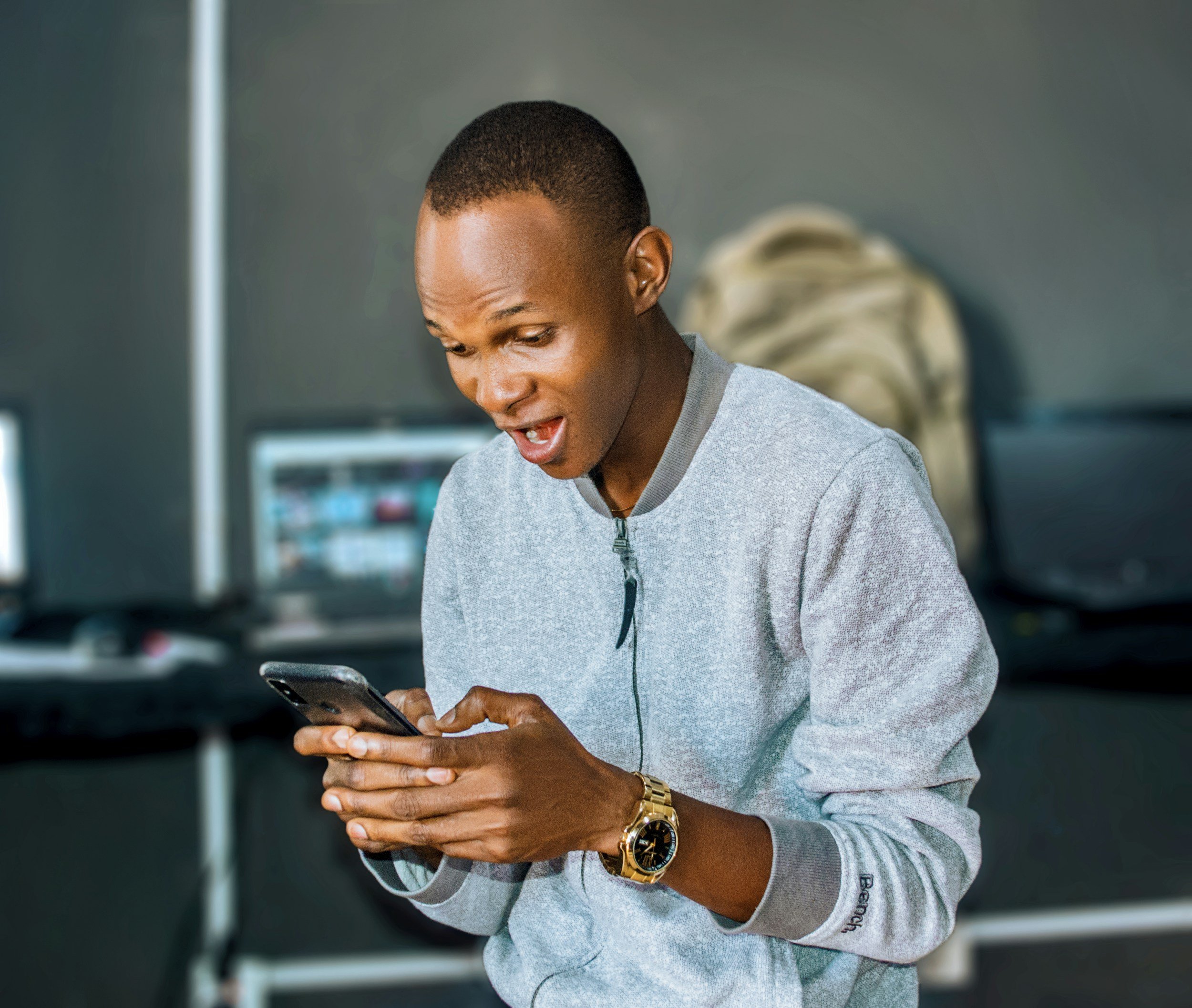 Young man in gray sweatshirt looking surprised while using a smartphone indoors.