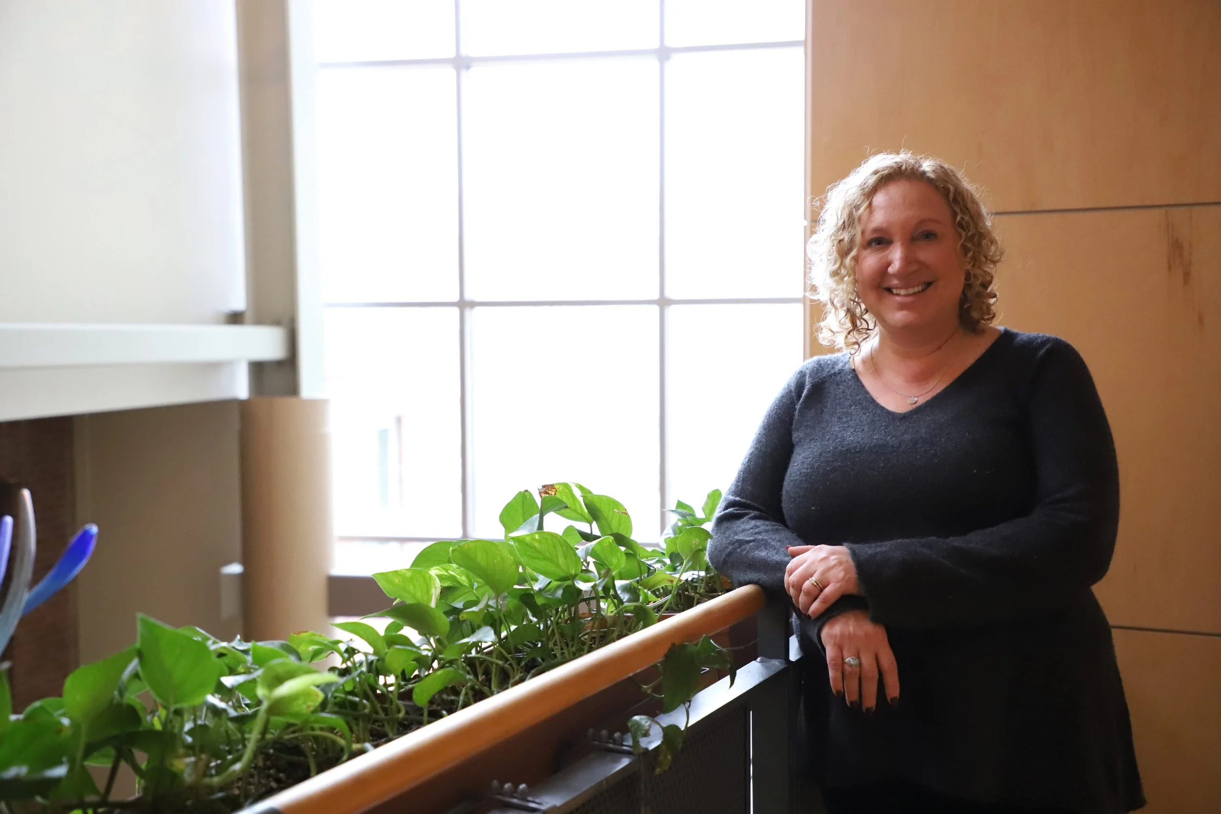 A woman with curly blonde hair smiling, standing indoors next to a window with sunlight, leaning on a planter box filled with green leafy plants.