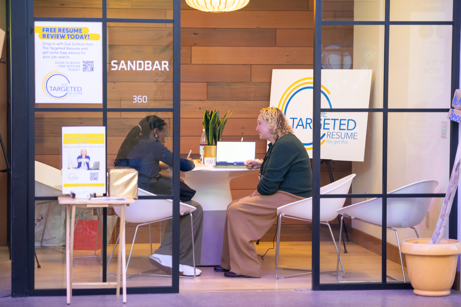 Two women sitting at a table inside a room labeled "SANDBAR" with a wooden wall background, a large "The Targeted Resume" sign, and promotional posters on the glass window.