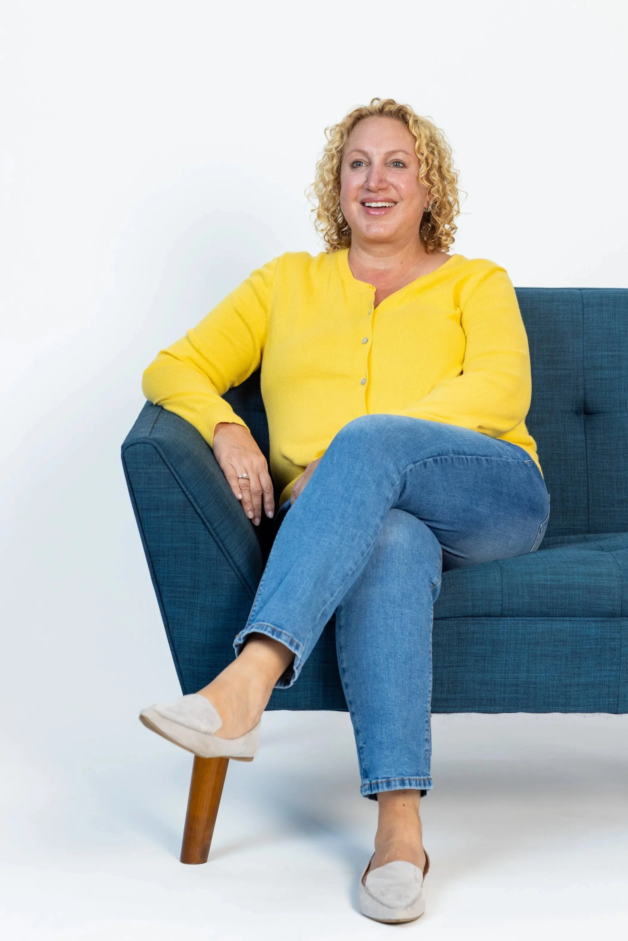 A woman with curly blonde hair sitting on a blue sofa. She is smiling, wearing a yellow cardigan, blue jeans, and beige shoes, against a plain white background.