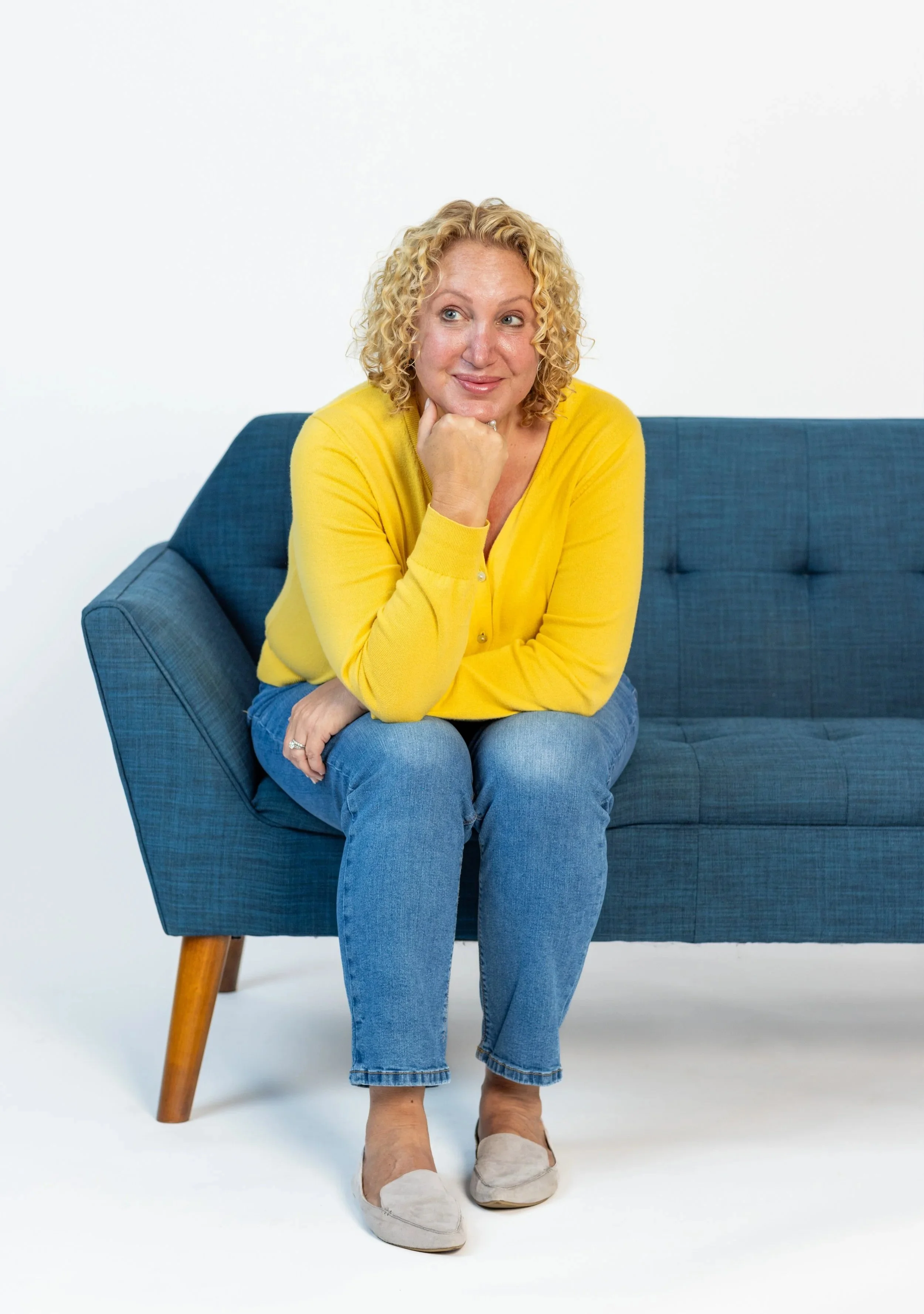 Woman with curly blonde hair wearing a yellow sweater and blue jeans sitting on a blue couch with one fist under her chin, looking to the side.