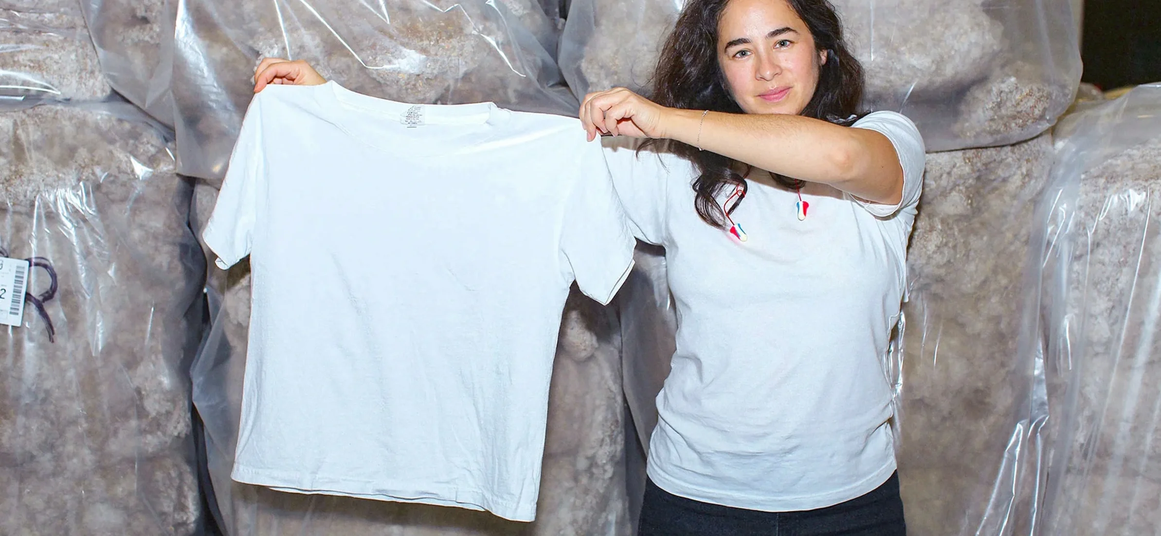 Woman holding up a sustainable t-shirt from Everybody World with large clear bags of reclaimed waste cotton visible in the background, demonstrating transparent sustainable production.