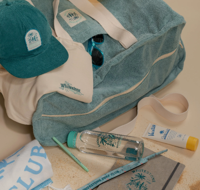 Beach essentials including a teal cap, a white towel, sunglasses, a blue duffel bag, a tube of sunscreen, a water bottle, a toothbrush, and a towel with