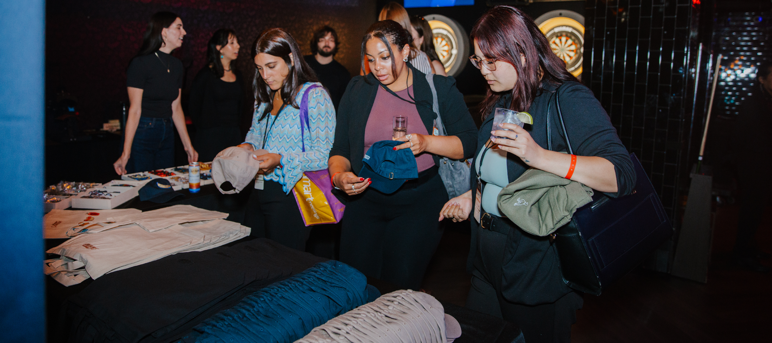 3 individuals selecting blank hats and tote bags in khaki and navy to move down an assembly line and decorate with custom patches.
