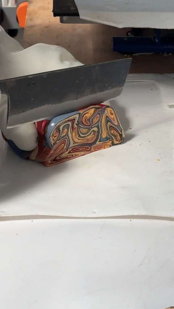 A close-up video of a worker wearing thick, white protective gloves using a large blade to slice through a warm, malleable log of melted recycled plastic, revealing a complex marbled interior pattern of blue, red, and yellow swirls.