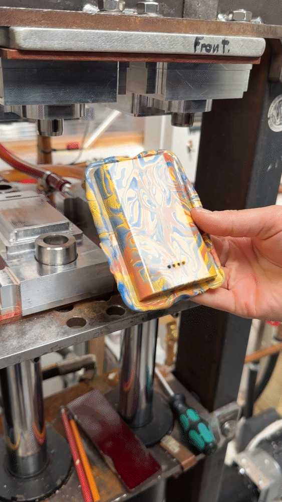 A close-up of a hand holding a newly formed, rectangular power bank shell with a bright yellow and blue marbled pattern as it is pulled from a heavy industrial metal compression press machine.