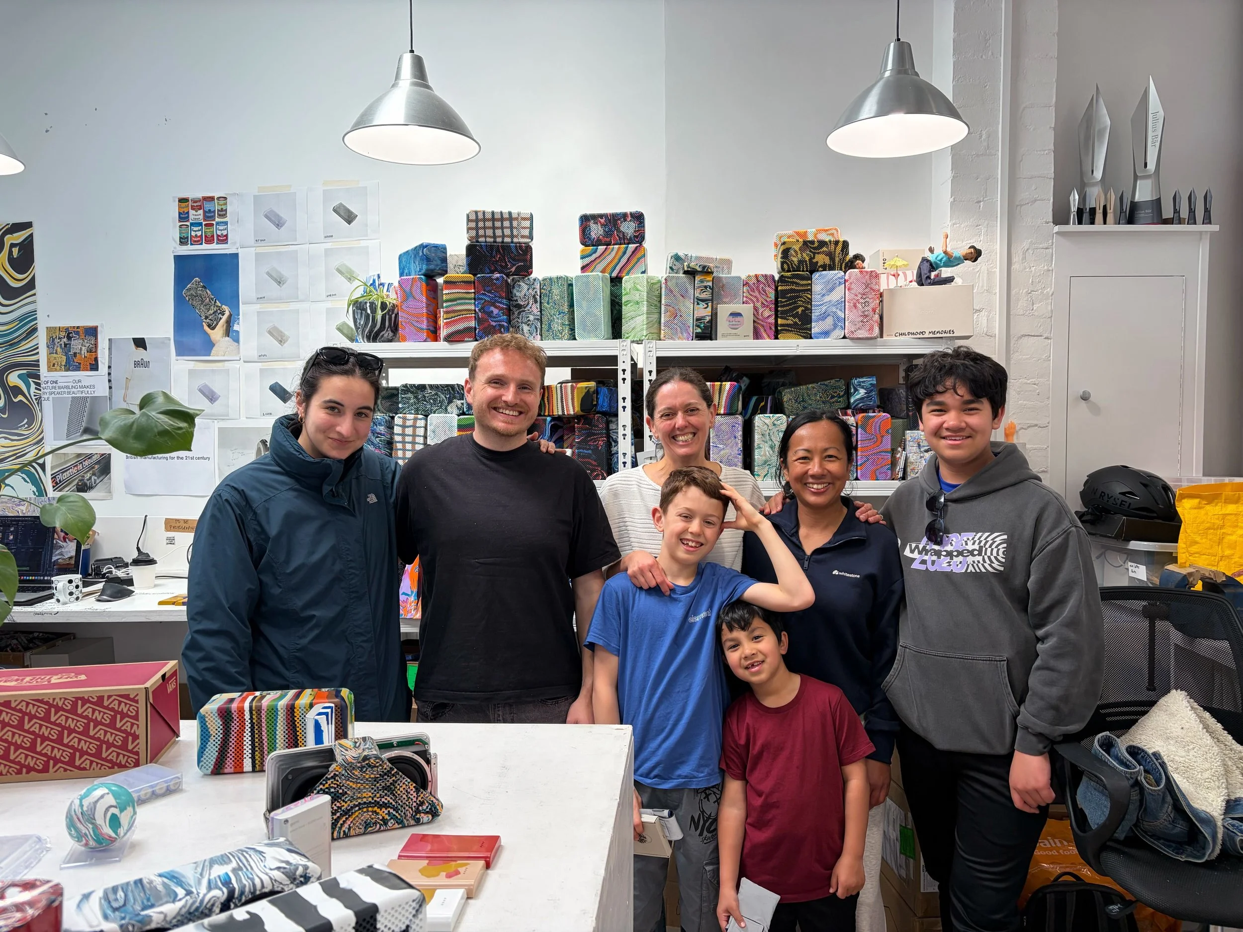 A group photo of the Whitestone and gomi teams, including two adults and three kids, smiling together inside the bright Gomi design studio surrounded by colorful recycled tech products and shelving.