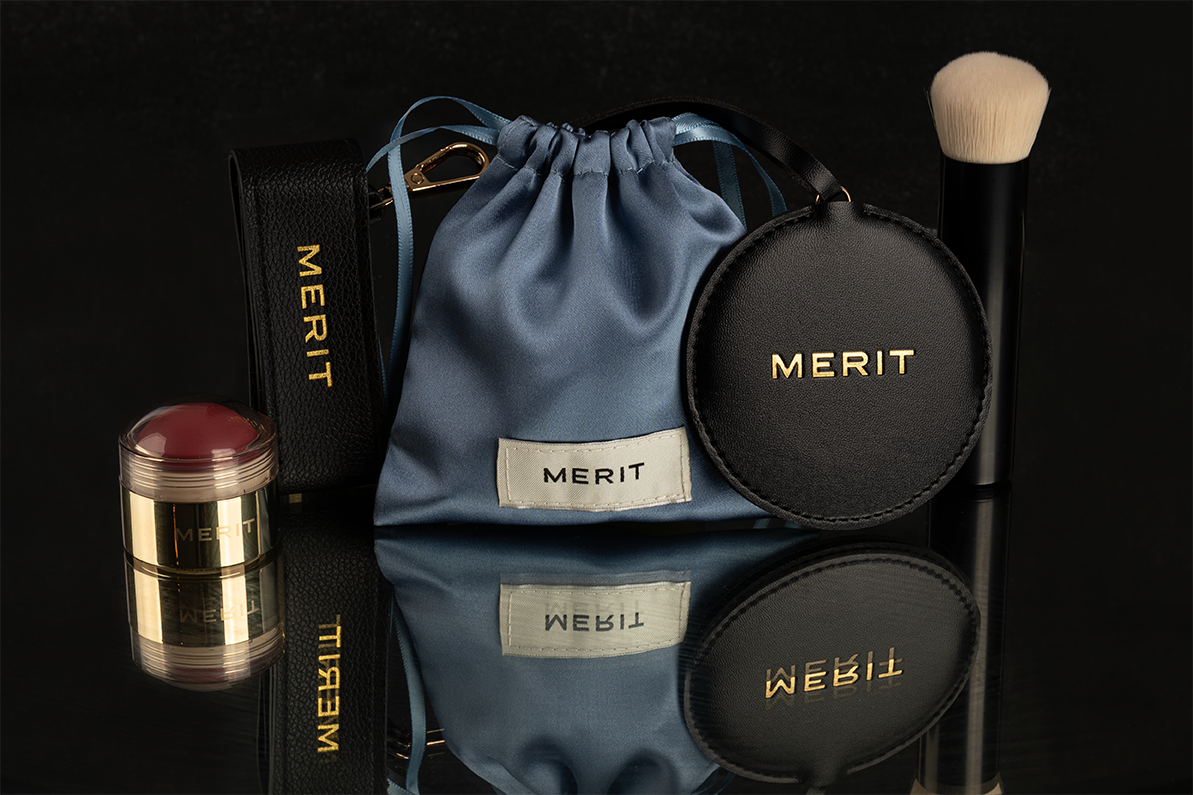 Collection of Merit cosmetics and makeup items, including a makeup brush, makeup bag, a compact, a compact mirror, and a beauty sponge, all arranged on a reflective black surface.