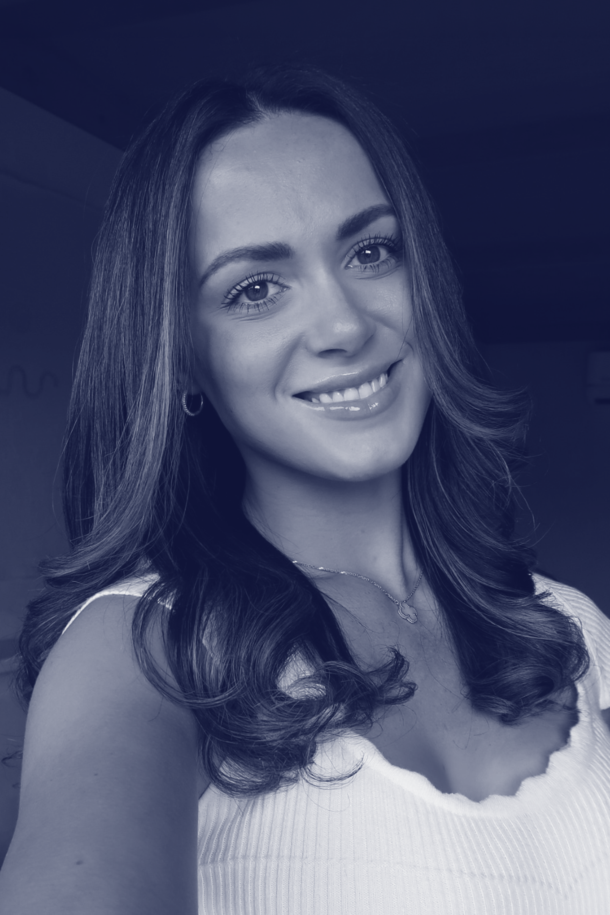 A woman with long, wavy hair smiling at the camera, wearing a white sleeveless top and a necklace, with a dark background.