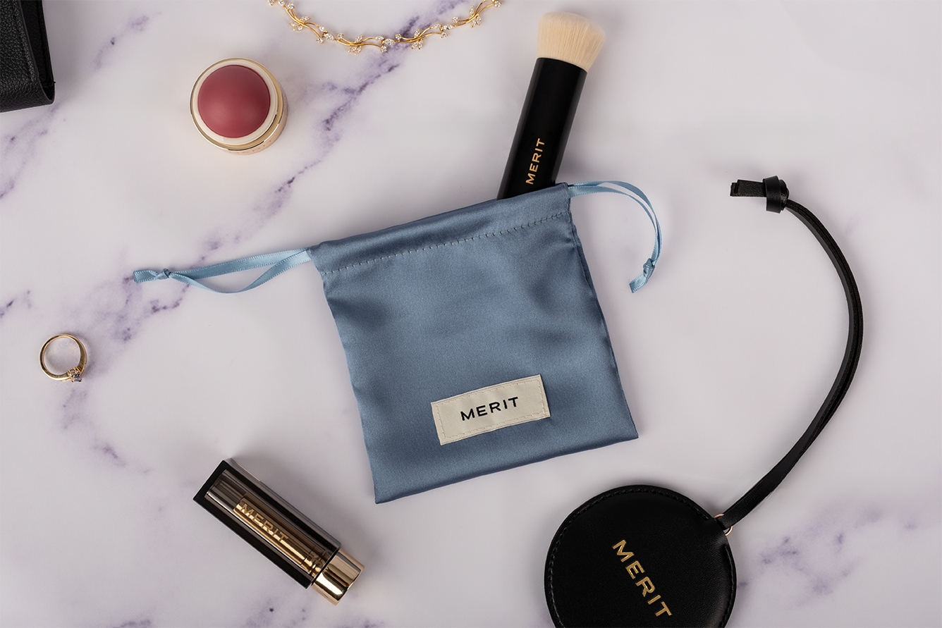 Flat lay of beauty and fashion accessories on a white marble surface, including a small blue Merit makeup pouch, a Merit face brush, a round lipstick, a black Merit round purse with a strap, a gold ring, a gold and pearl necklace, and a black container in the top left corner.
