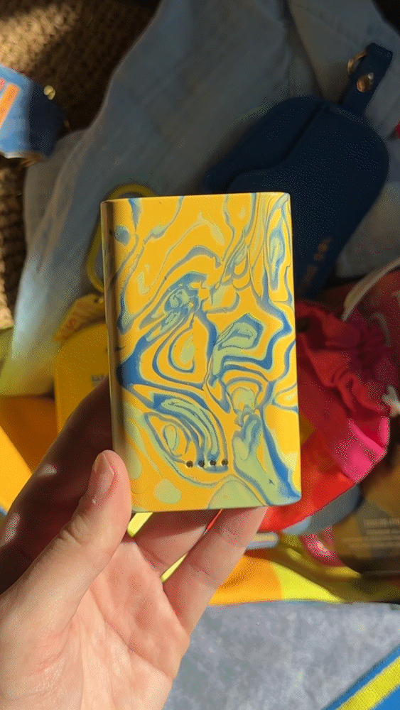 A close-up GIF of a person holding a rectangular Gomi power bank from the Sol Collection. The power bank features a vibrant, hand-marbled swirl pattern of bright yellow, ocean blue, and pale green, made from 100% recycled plastic waste.