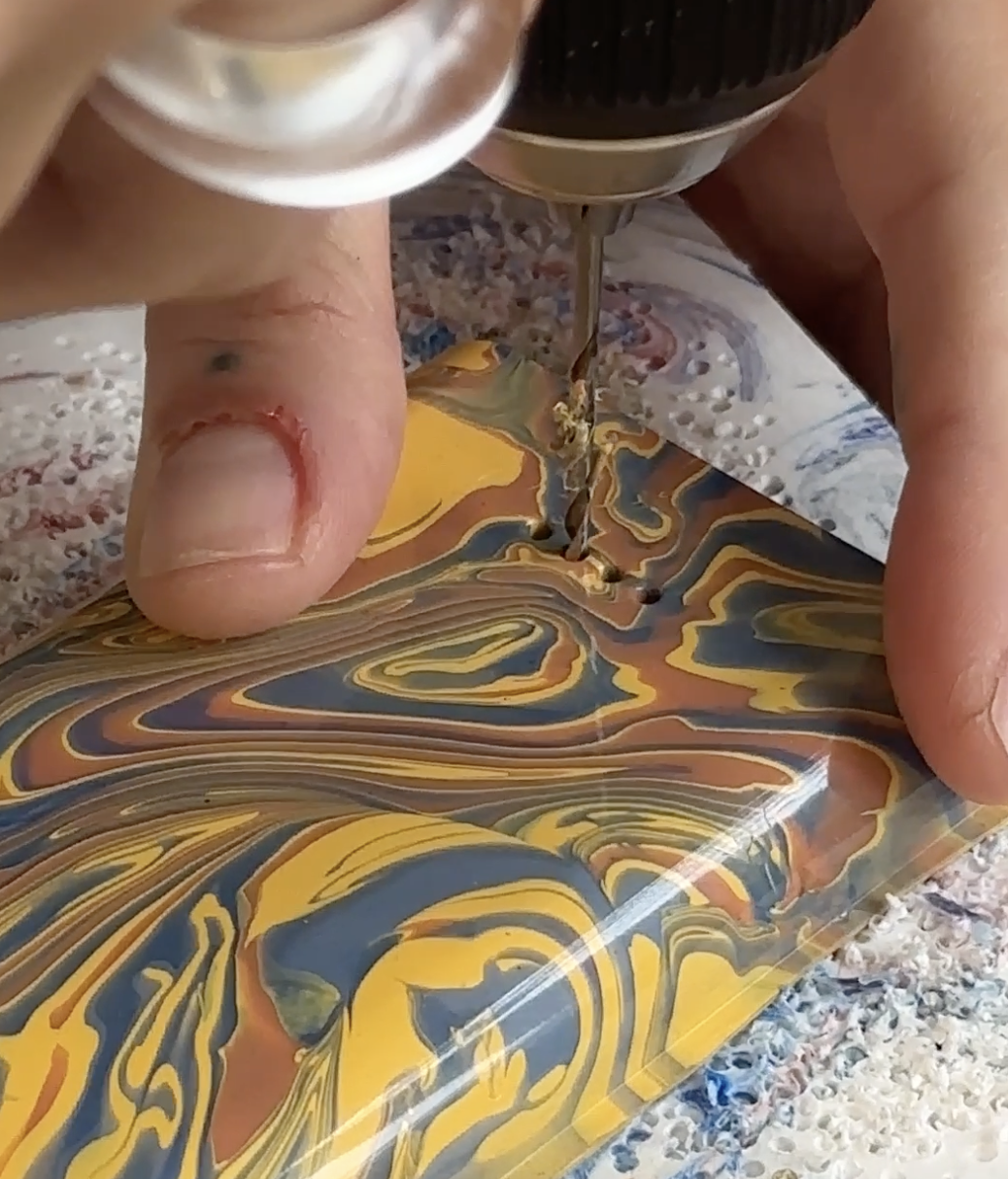 A close-up of a worker's hands using a precision drill bit to carefully create a small hole in a glossy, marbled orange, yellow, and blue recycled plastic power bank casing.