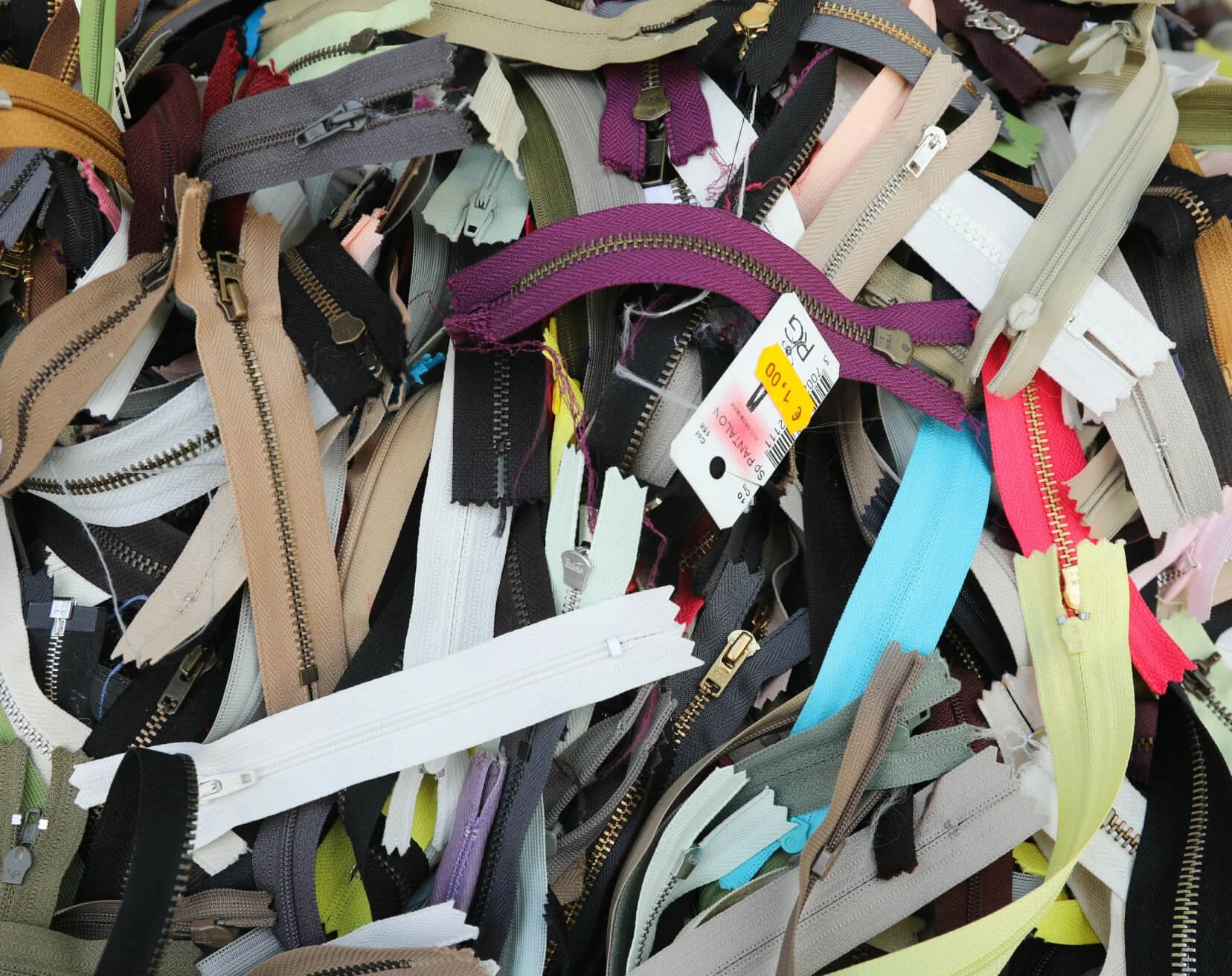 A detailed top-down close-up shot of a tangled heap of salvaged zippers in various colors, styles, and materials, including plastic and metal teeth, with some still bearing tags.