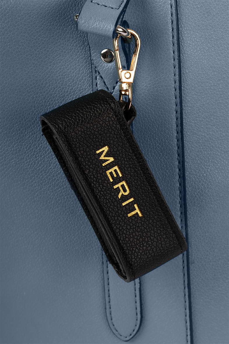 A close-up of a blue textured leather bag with a black leather key holder that has the word "MERIT" in gold letters, attached to a metal hook.