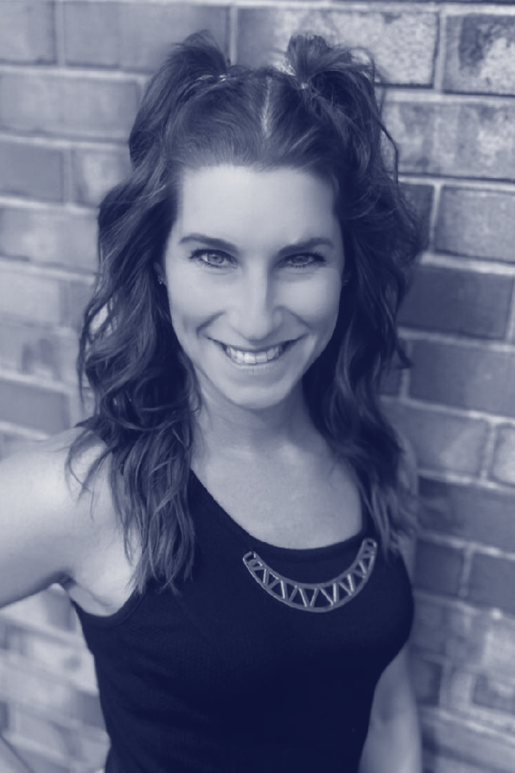 A smiling woman with wavy hair styled in pigtails, wearing a sleeveless black top and a statement necklace, standing in front of a brick wall.