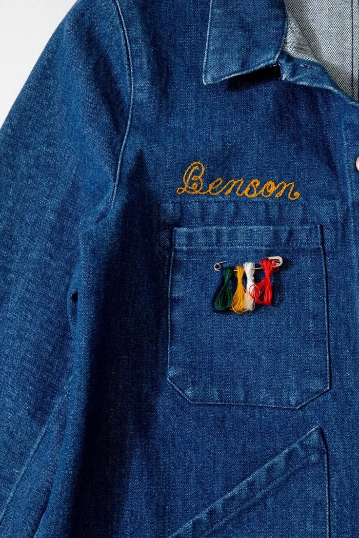Vintage denim jacket featuring custom embroidered name above the chest pocket with a colorful yarn pin attached, showing creative reuse and personalization.