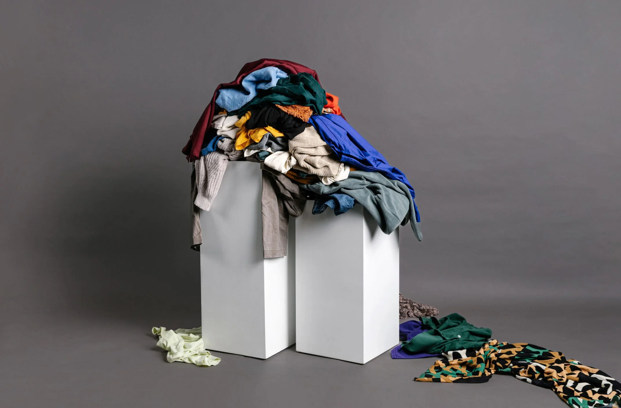 A studio photo featuring a large, disorganized mound of colorful discarded clothing piled on top of two minimalist white rectangular columns against a solid grey background.