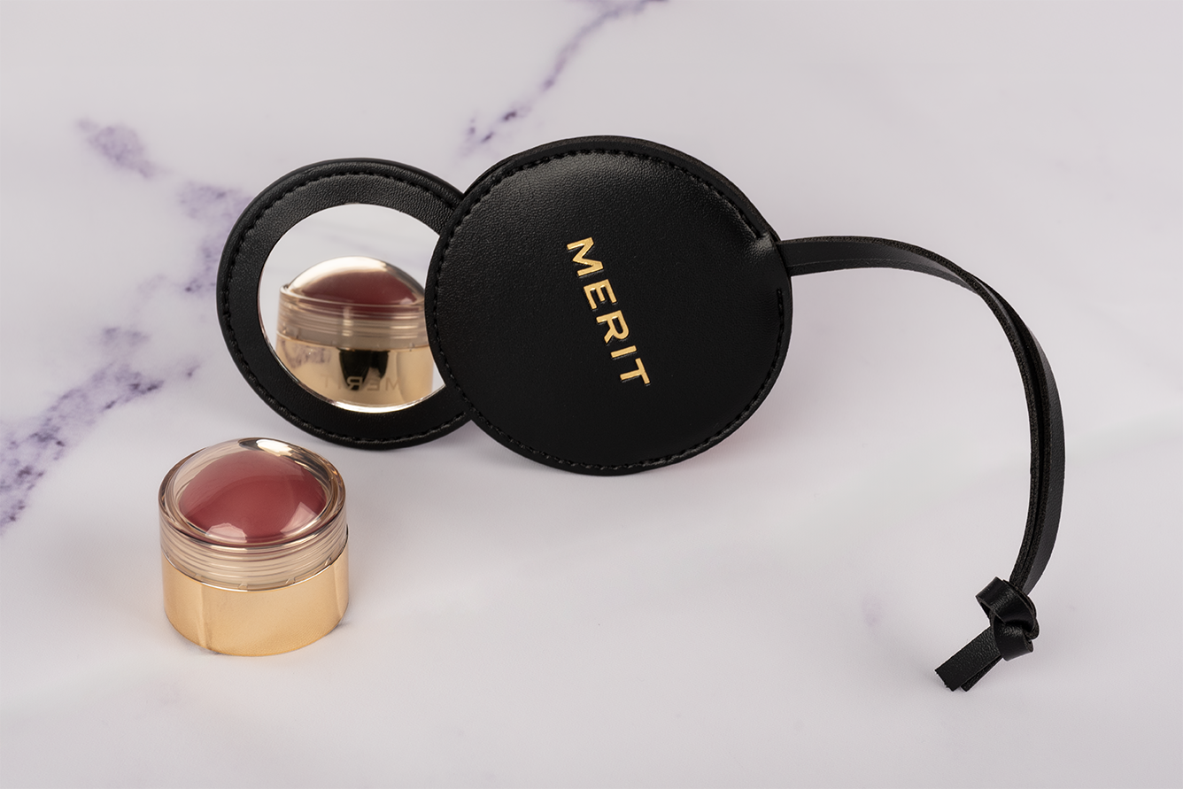 Cosmetic product with a round container and a mirror, along with a black circular case labeled 'MERIT' on a white marble surface.