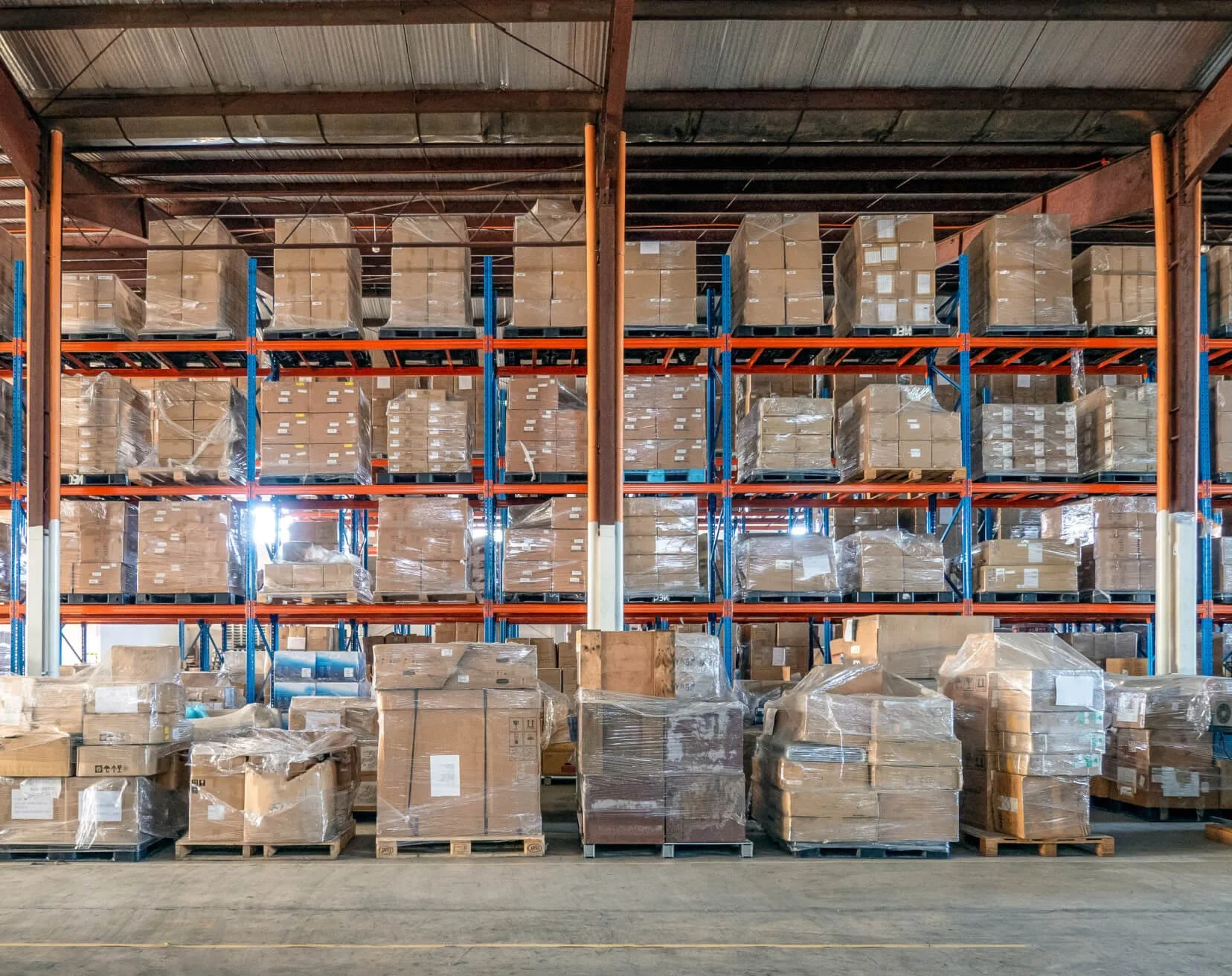 A wide shot of a massive industrial logistics warehouse showing tall, multi-level orange and blue pallet racks loaded with shrink-wrapped cardboard boxes, alongside staged pallets on the concrete floor.