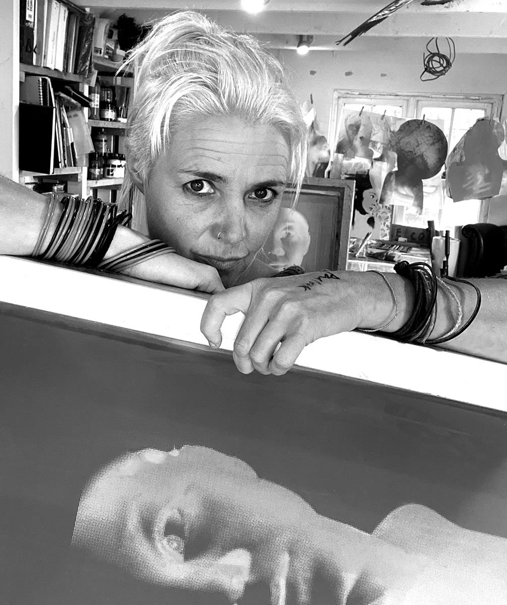 A woman with light-colored hair resting her arm on a surface, looking intently at the camera. The background includes bookshelves, art supplies, and hanging paper mache or papier-mâché objects. In the foreground, an illuminated monitor displays a digital portrait of a face.