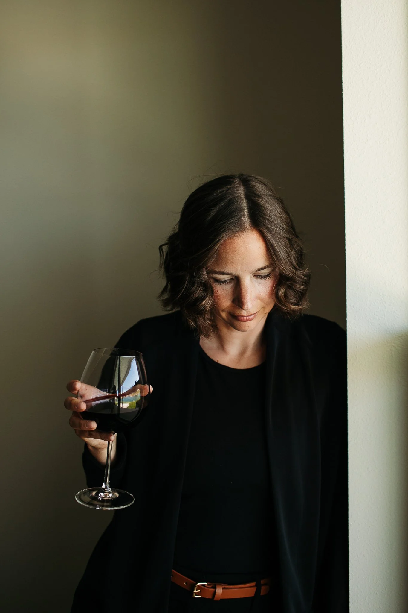 Woman with wavy brown hair holding a glass of red wine, standing near a wall, looking down, wearing a black top and jacket.