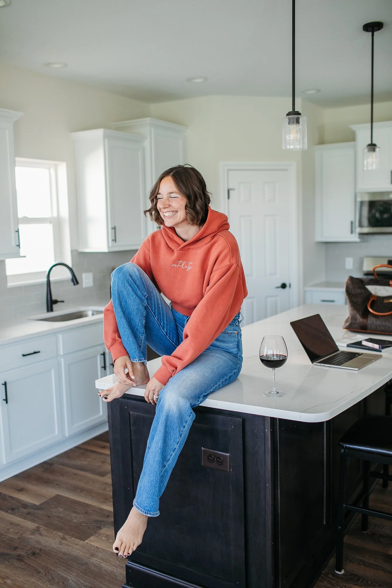 A woman in a red hoodie and blue jeans smiling and sitting on a kitchen island, holding her foot. There is a glass of red wine, a laptop, and a bag on the counter.