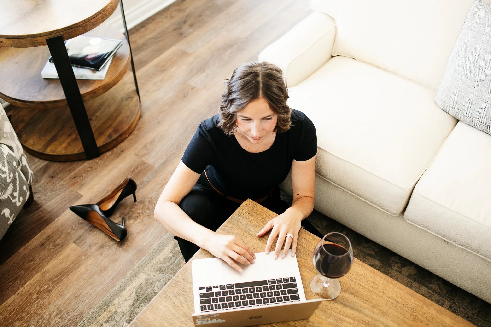 A woman sitting on the floor in a living room, working on a laptop with a glass of red wine nearby.