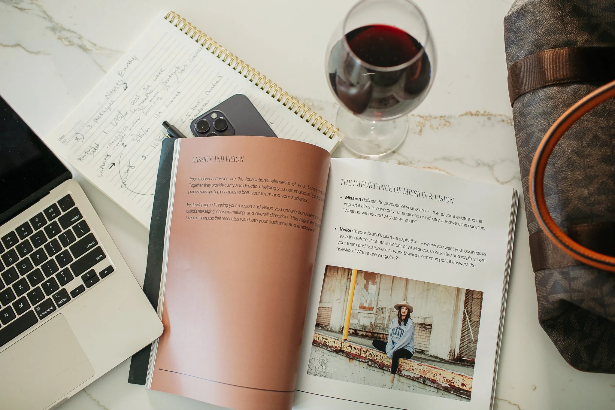 A workspace with a MacBook laptop, an open notebook with handwritten notes, an iPhone, a glass of red wine, an open book about mission and vision with a picture of a woman sitting on a ledge, and a Louis Vuitton bag on a marble surface.