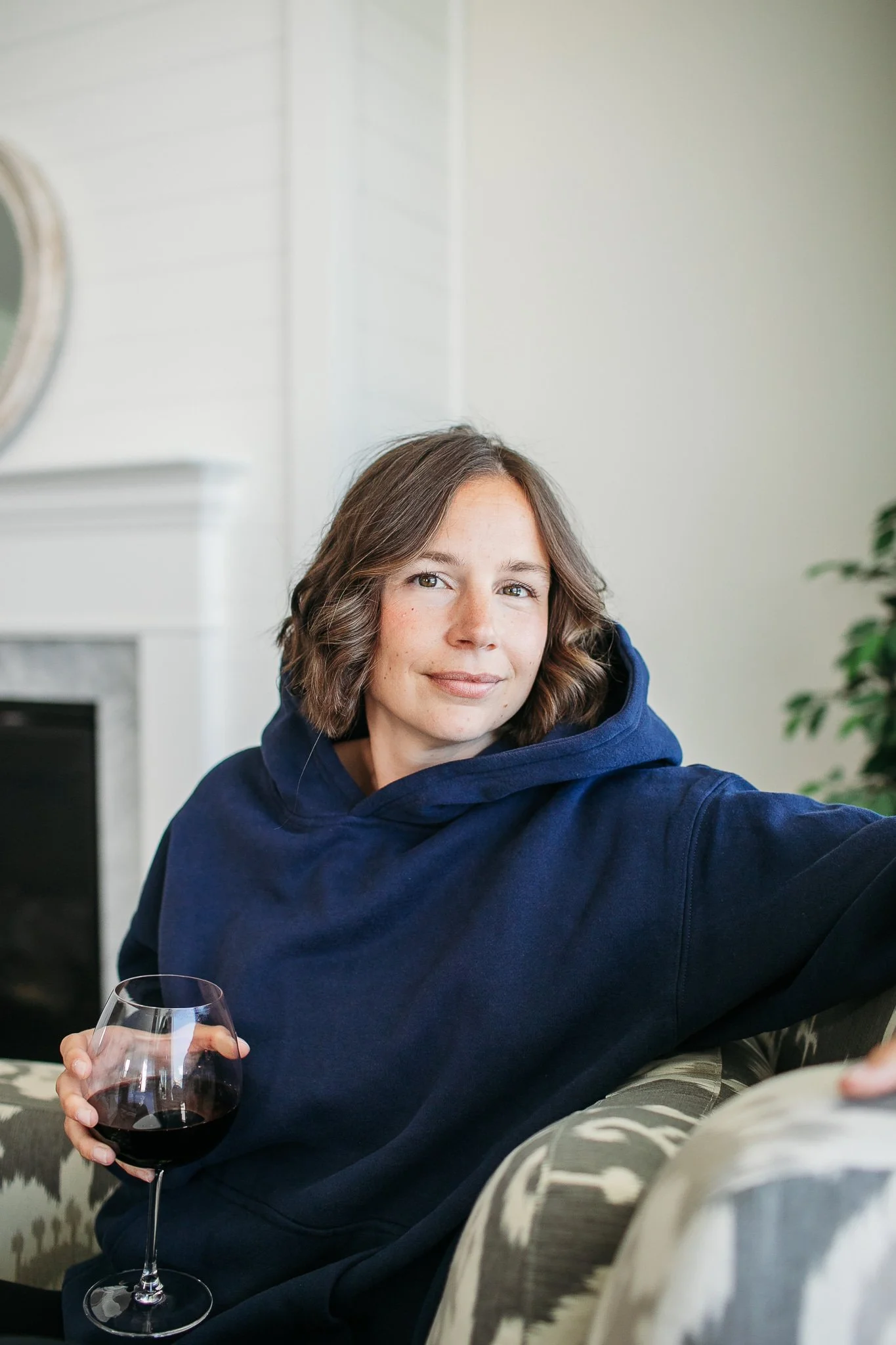 A woman with short brown hair, wearing a navy blue hoodie, sitting on a patterned sofa, holding a glass of red wine in her right hand, smiling softly at the camera.