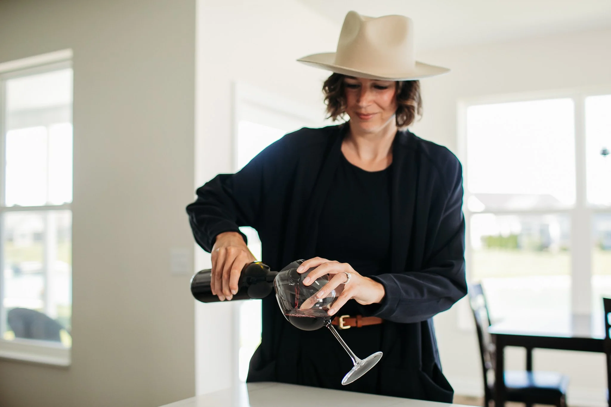 A woman wearing a beige wide-brimmed hat and black clothing pouring red wine into a glass in a bright, modern kitchen.