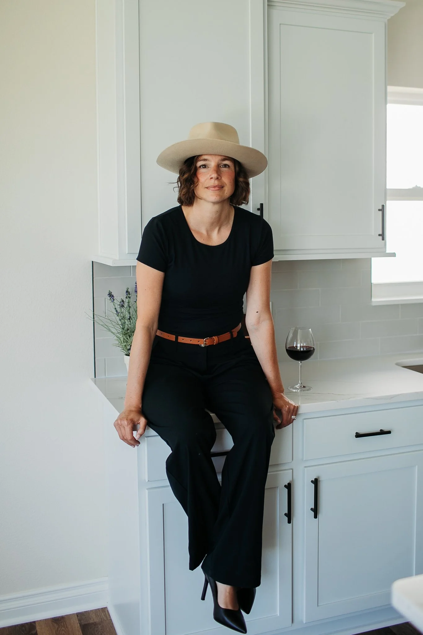 Woman sitting on kitchen counter with a glass of red wine, wearing a black outfit and beige hat, in a modern white kitchen.