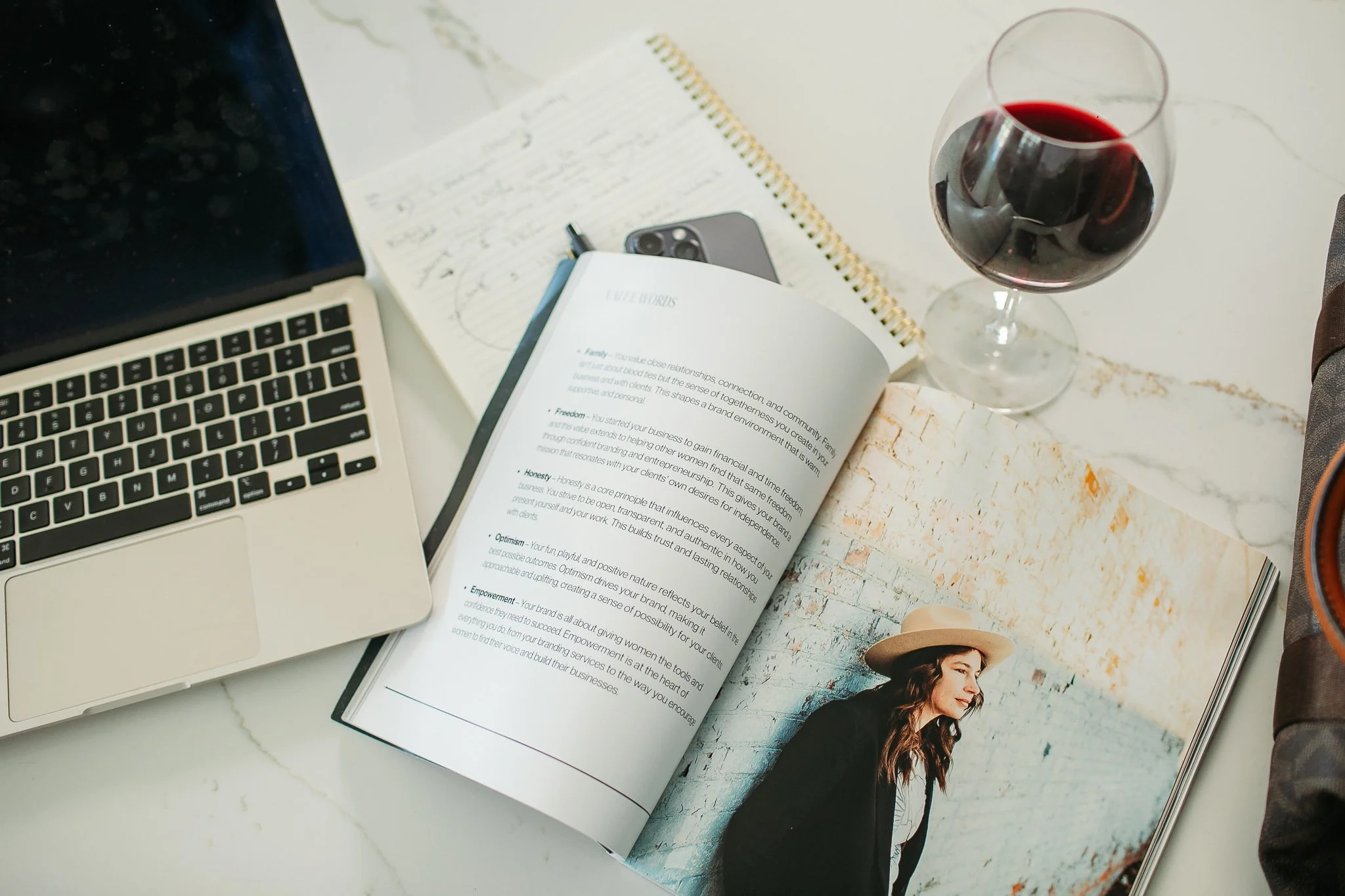 A workspace with a laptop, notebooks, a smartphone, a pen, a glass of red wine, and an open magazine showing a woman in a hat against a brick wall.