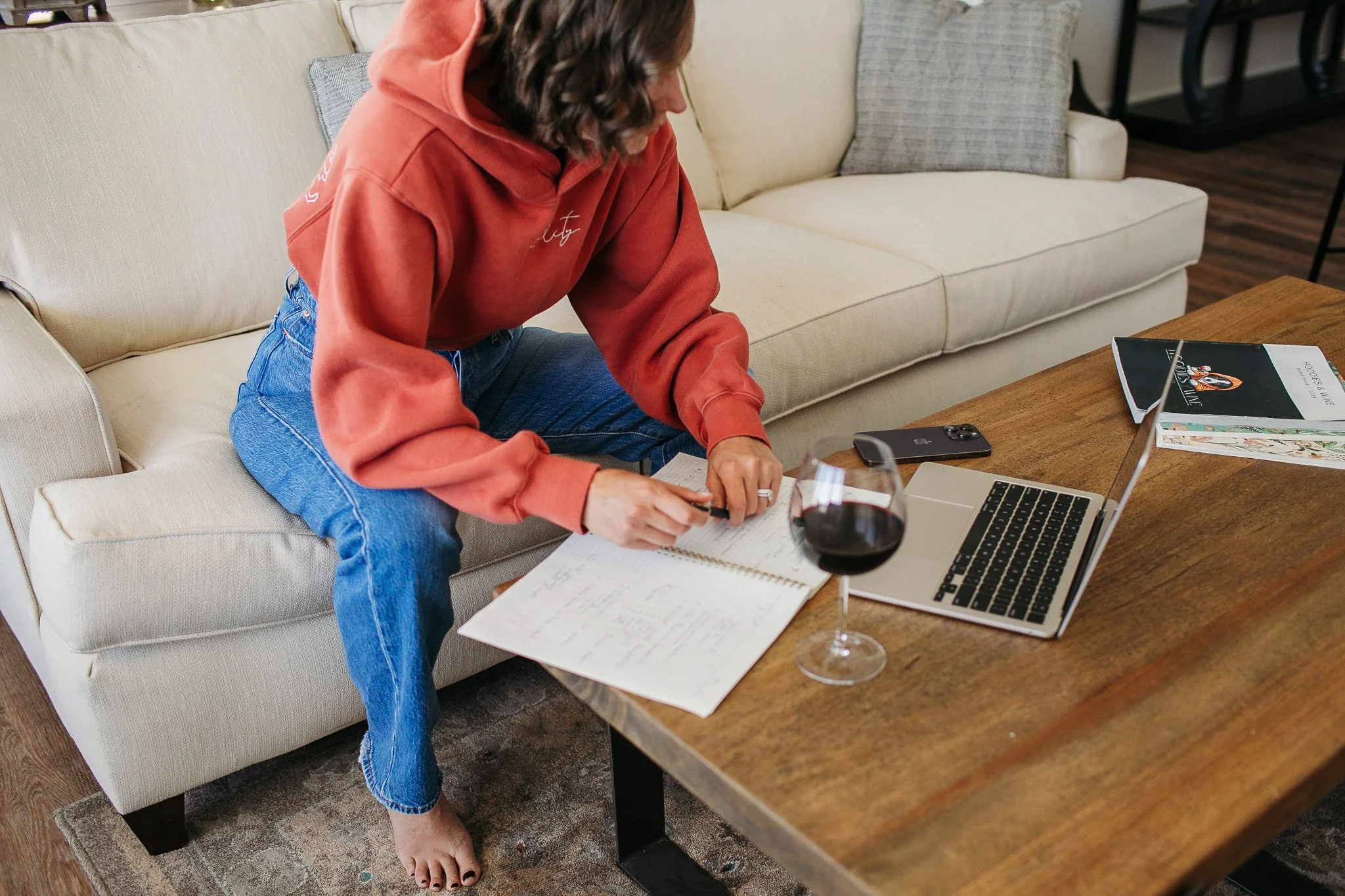 A woman in a red hoodie and blue jeans sitting on a beige couch, writing in a notebook on a wooden coffee table. There is a glass of red wine, a laptop, a smartphone, and a magazine on the table.