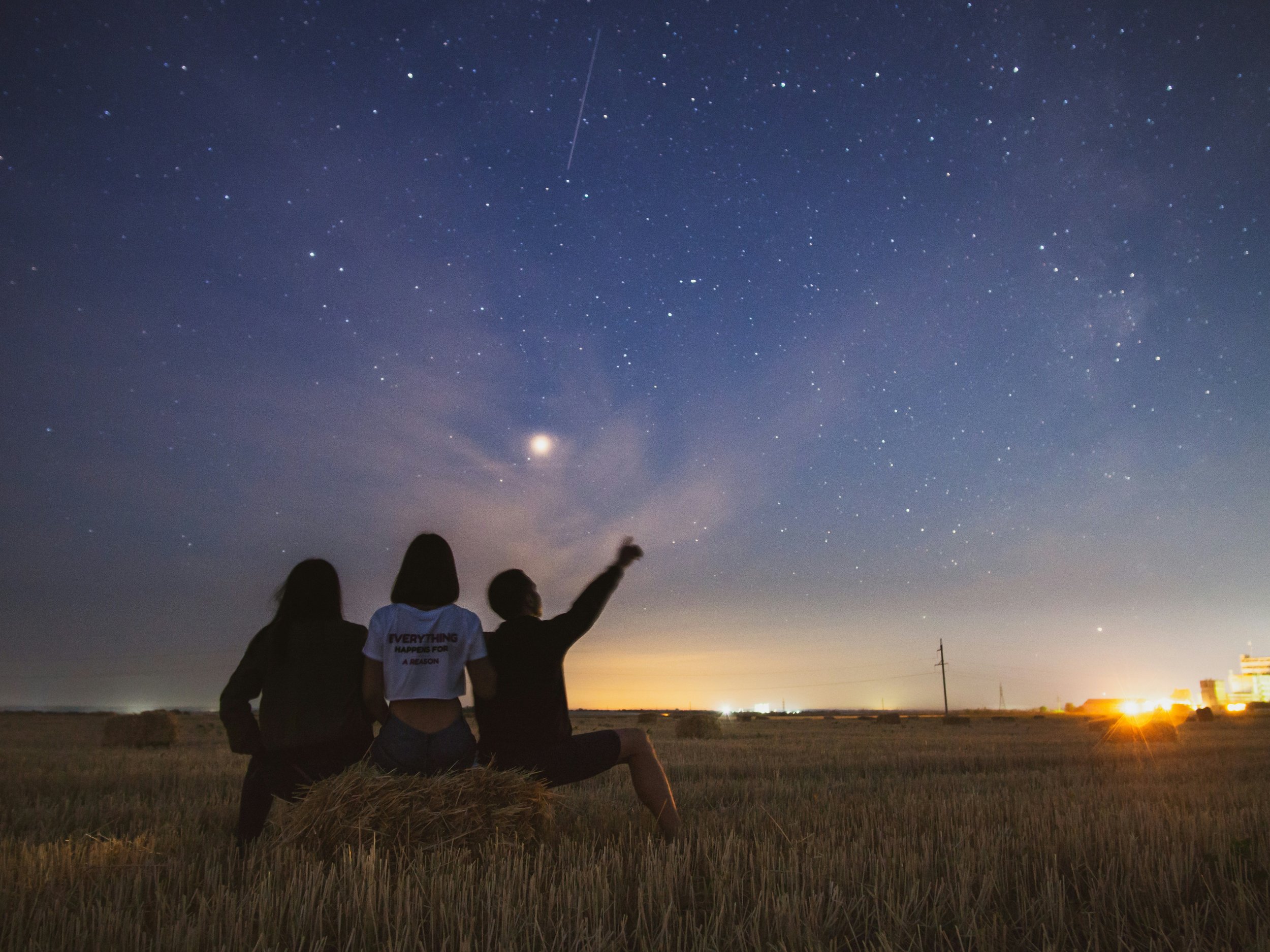 Three people sitting on a field at night, looking up at a starry sky and pointing towards a large planet or moon, with some distant lights on the horizon.