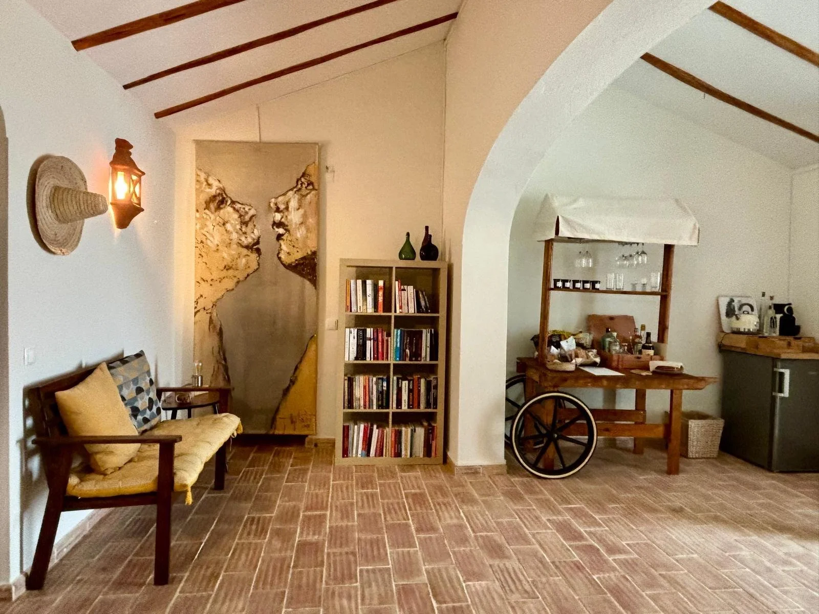 Living room with tiled floor, a wooden bench with cushions, a bookshelf filled with books, and a wooden cart with various items. There's a decorative wall hanging depicting a mountain scene, and a wall-mounted light fixture next to a straw hat on the wall. An arched opening leads into another area in the room.