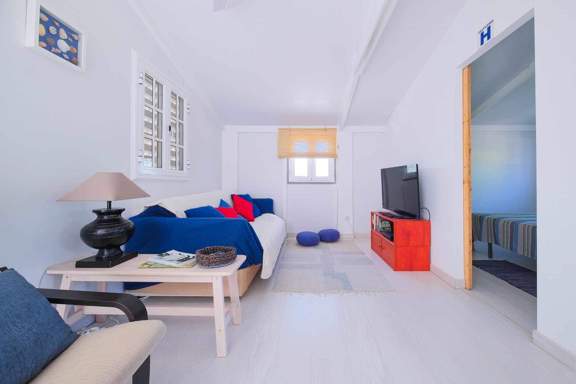 Living room with white walls, a white sofa with blue and red cushions, a wooden coffee table with a lamp and magazines, a television on a red stand, two blue floor cushions, and a window with blinds, leading to a bedroom with a striped bedspread.