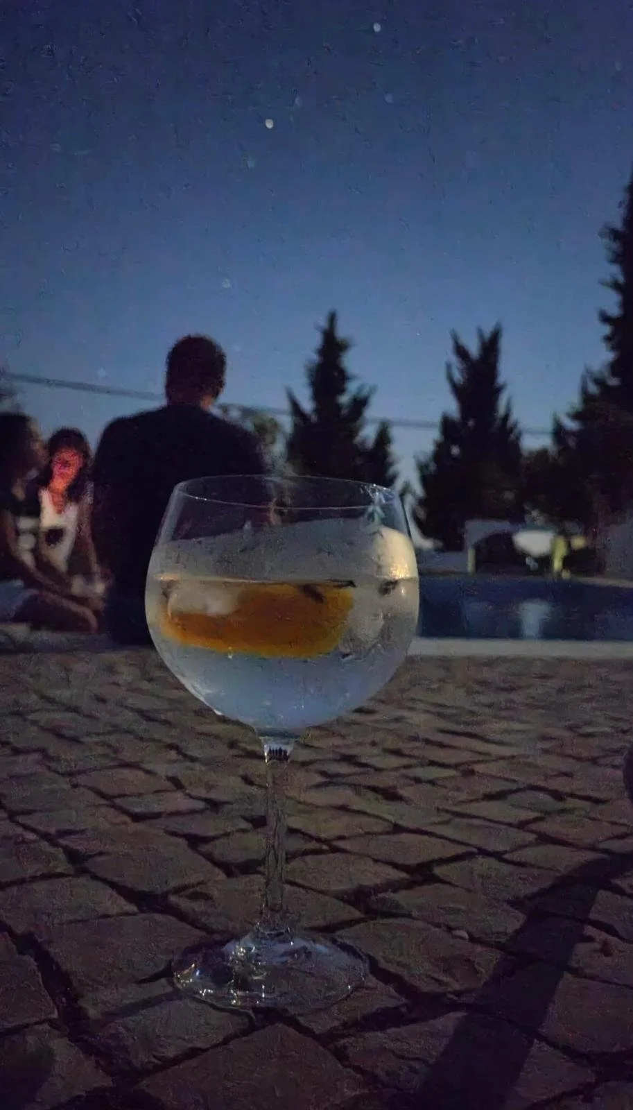 A clear cocktail glass with lemon slices and ice on a cobblestone surface outdoors at night, with people sitting and trees in the background under a starry sky.