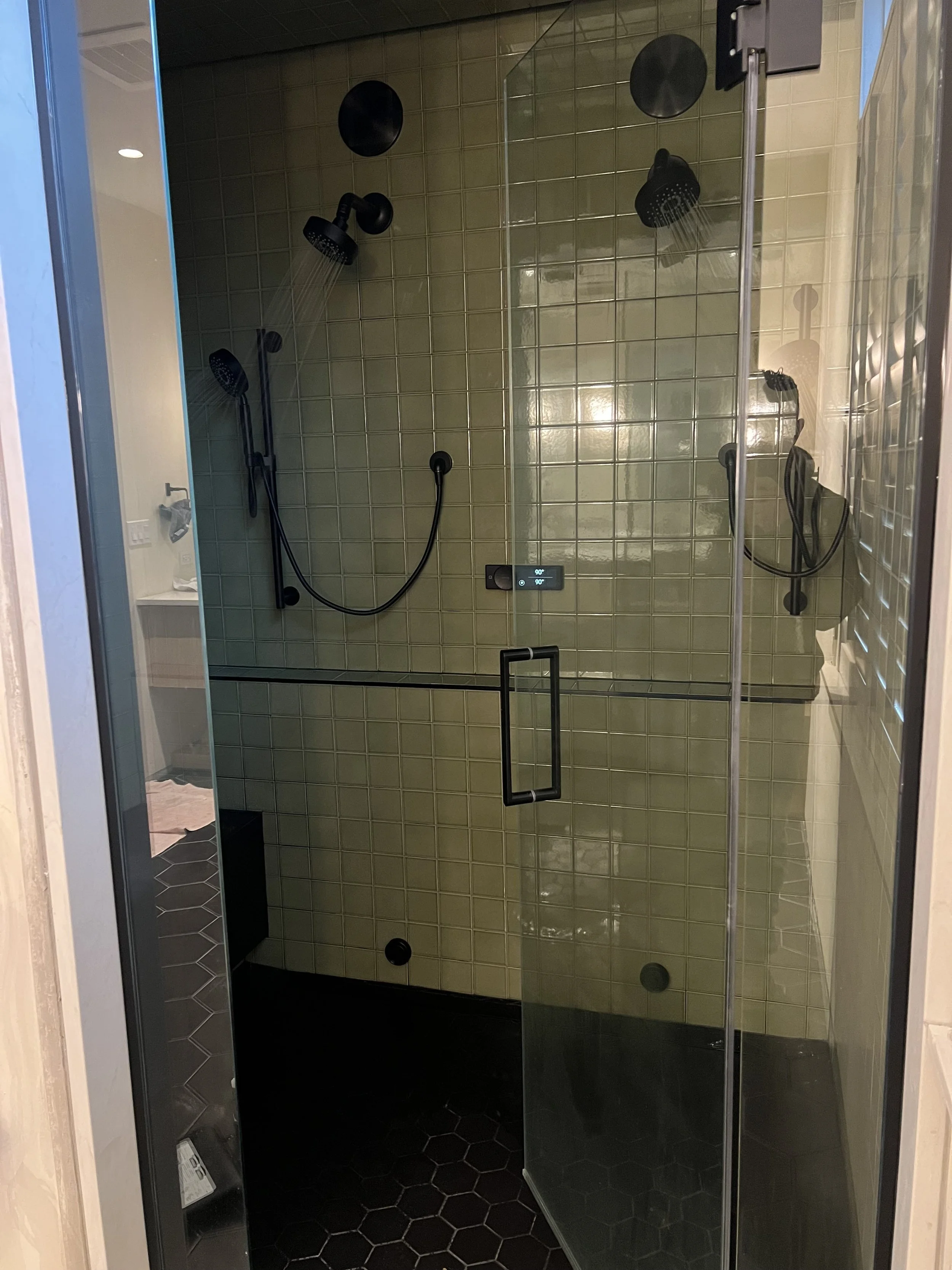 A glass-enclosed shower with green tiles on the wall, black fixtures, and multiple showerheads including a handheld. The shower has a digital control panel and black hexagonal floor tiles.