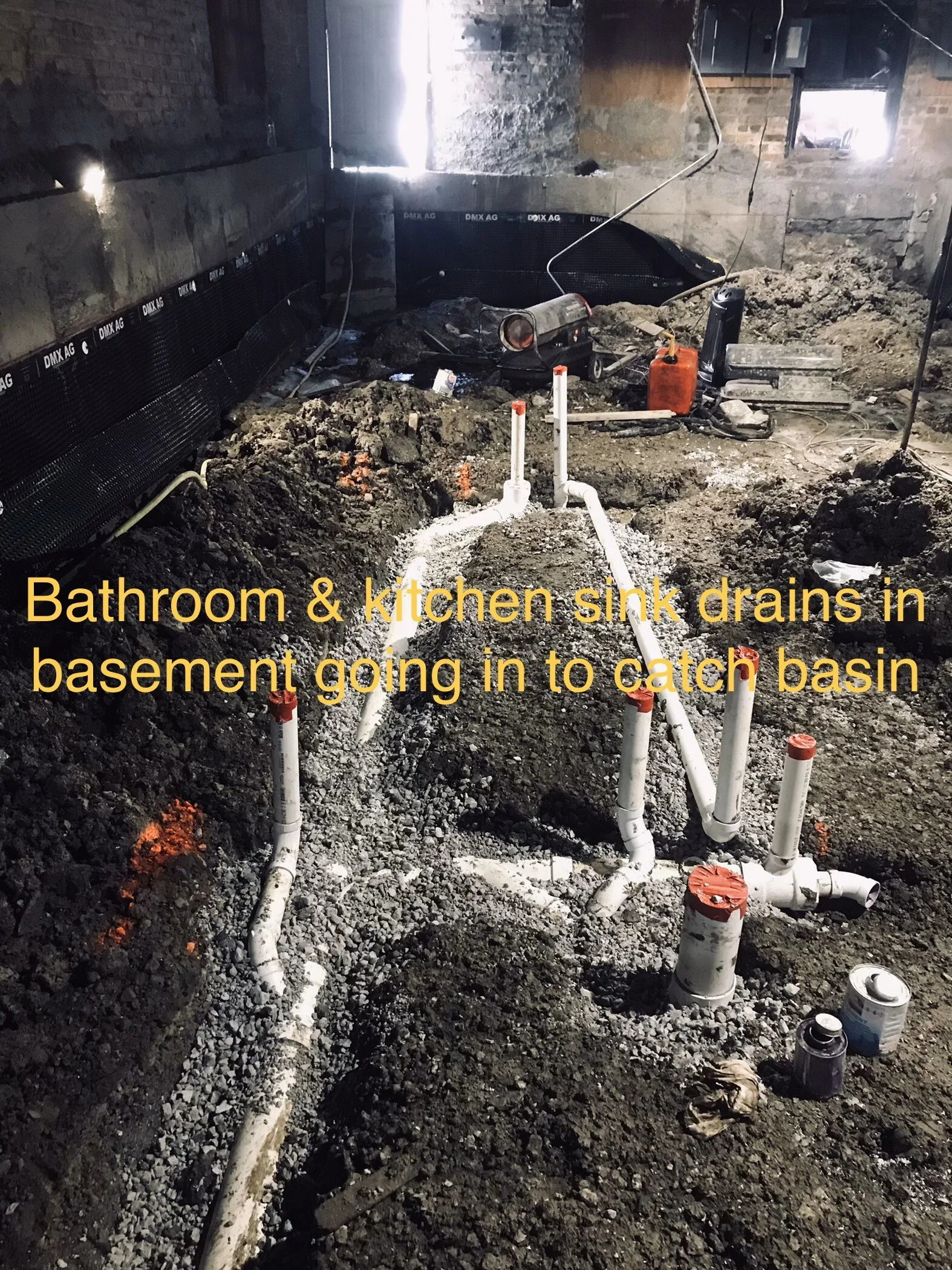 Construction site in a basement with exposed pipes, dirt, and tools, indicating ongoing plumbing work. Text overlay says: 'Bathroom & kitchen sink drains in basement going in to catch basin'.