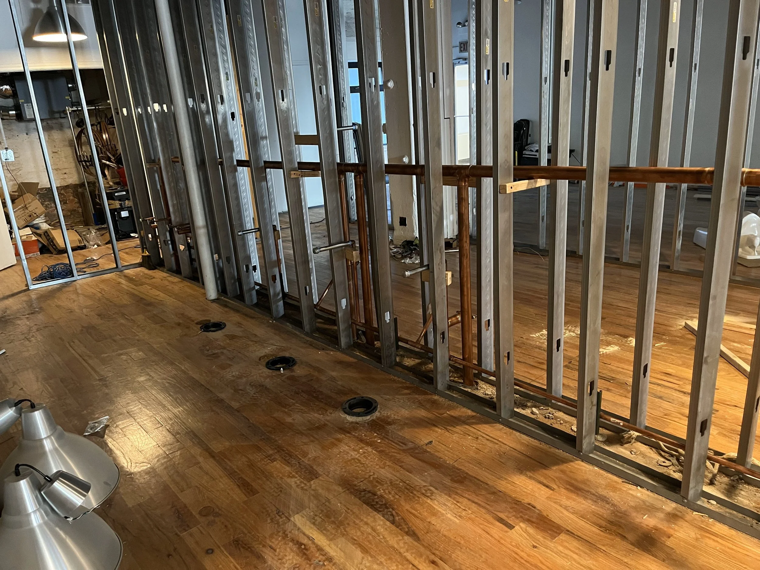 Interior of a commercial building under renovation with metal studs framing walls, a wooden handrail, and construction debris on the floor.