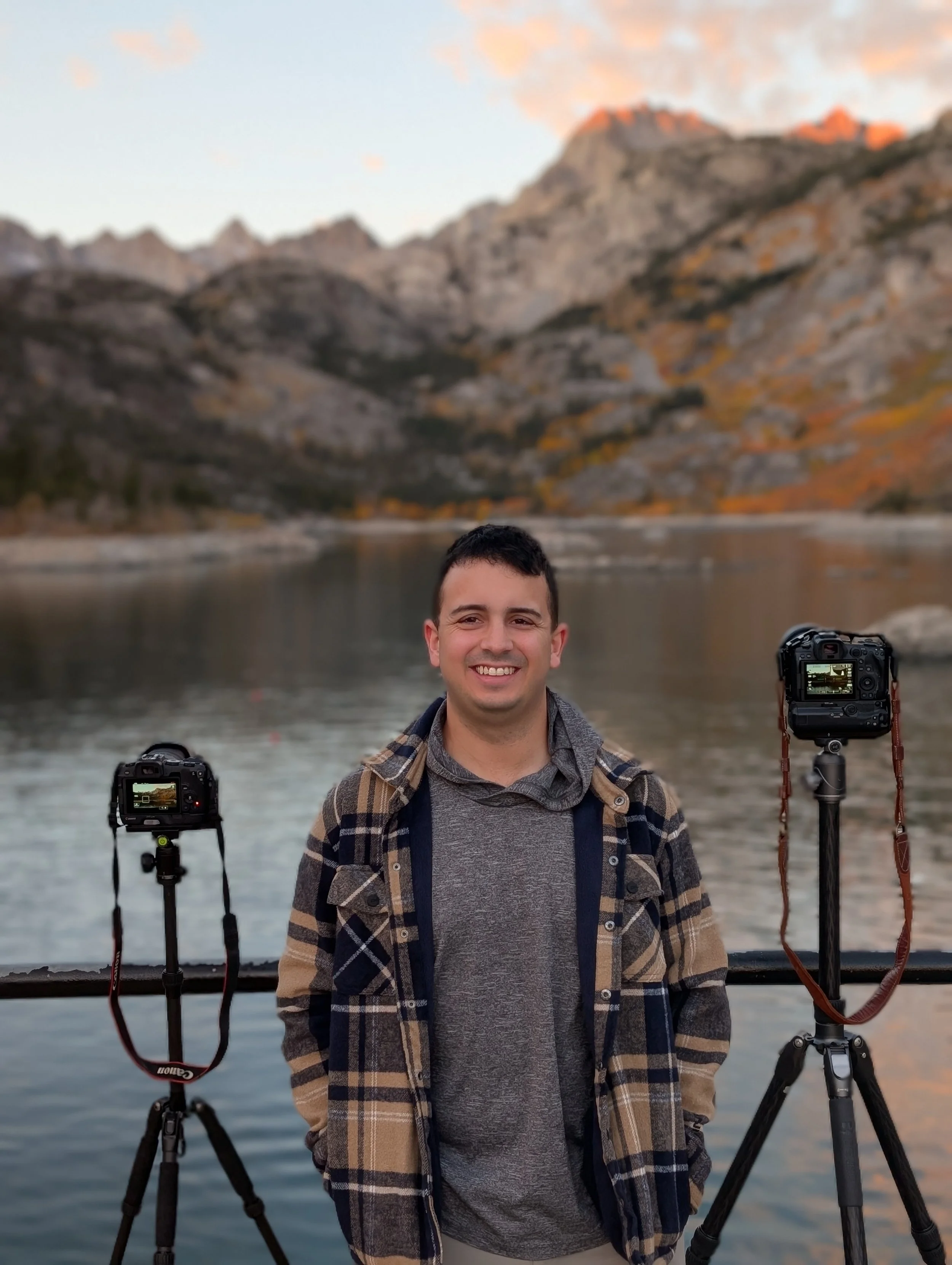 Smiling man wearing a flannel at sunrise with a mountain landscape, rocky terrain, and a camera on a tripod in the background.