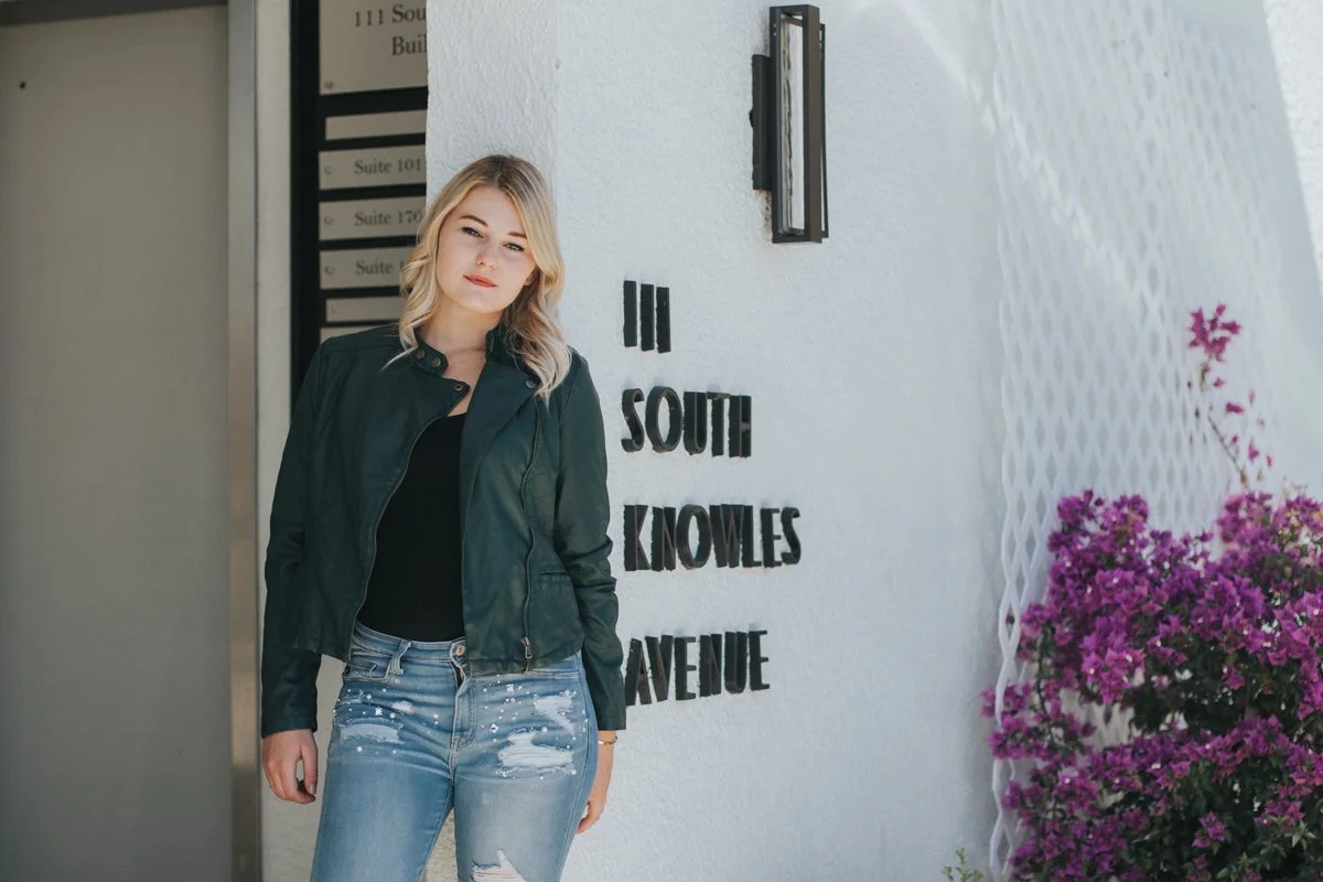 A young woman with blonde hair wearing a black leather jacket and ripped jeans standing outside a white building with a sign that reads '111 South Knowles Avenue' and vibrant pink flowers to the right.