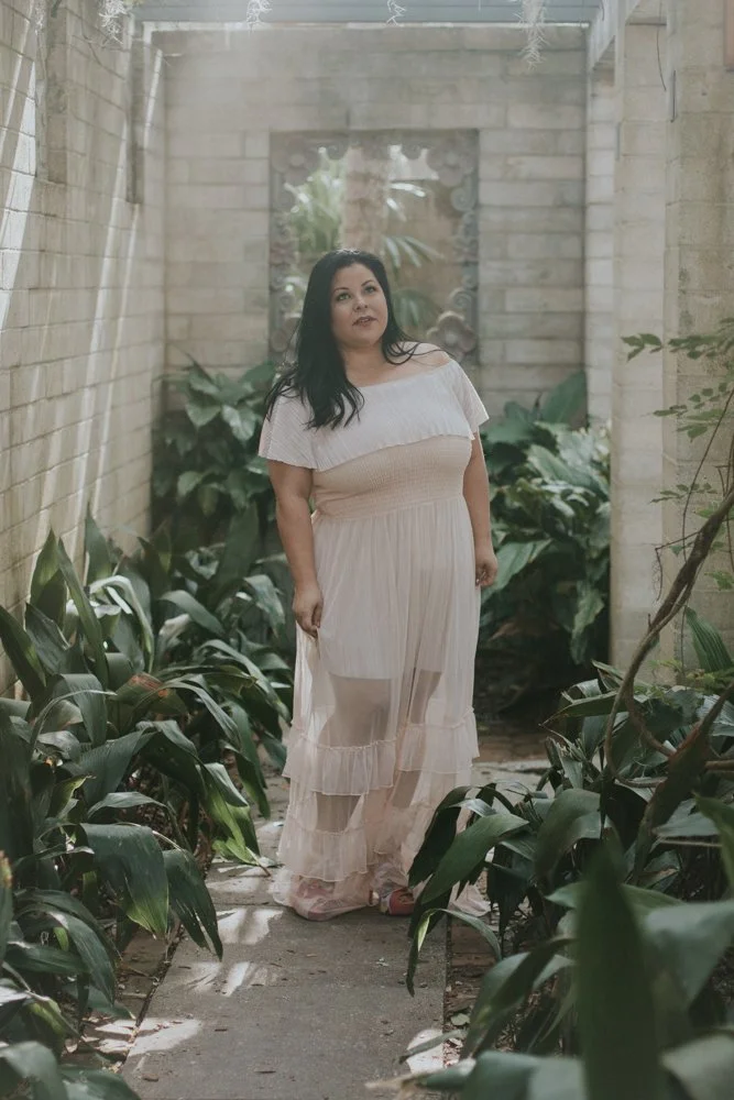A woman in a light, flowing off-shoulder dress stands on a narrow garden path surrounded by green plants, with a brick wall and a small window in the background.