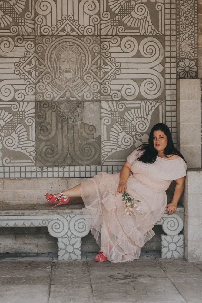 A woman in a pale pink dress and red floral shoes reclining on a stone bench in front of a carved stone wall with religious and decorative patterns, holding a small bouquet of flowers.