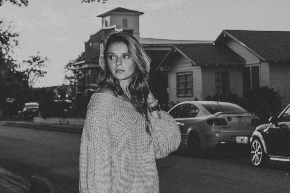 A young woman with wavy hair wearing an oversized sweater stands on a suburban street at dusk, with houses and parked cars behind her.