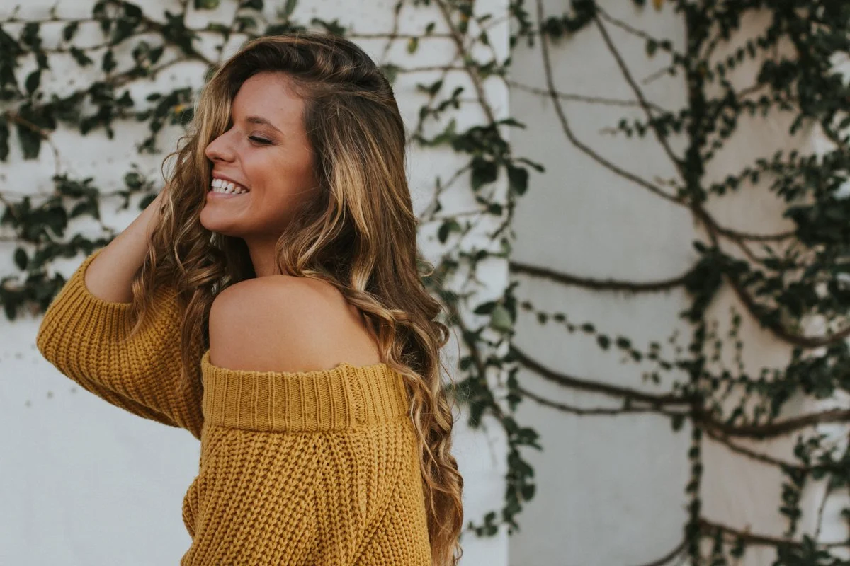 A woman with long, wavy hair smiling and posing with her eyes closed, wearing a yellow off-the-shoulder sweater, standing outdoors against a wall with creeping vines.