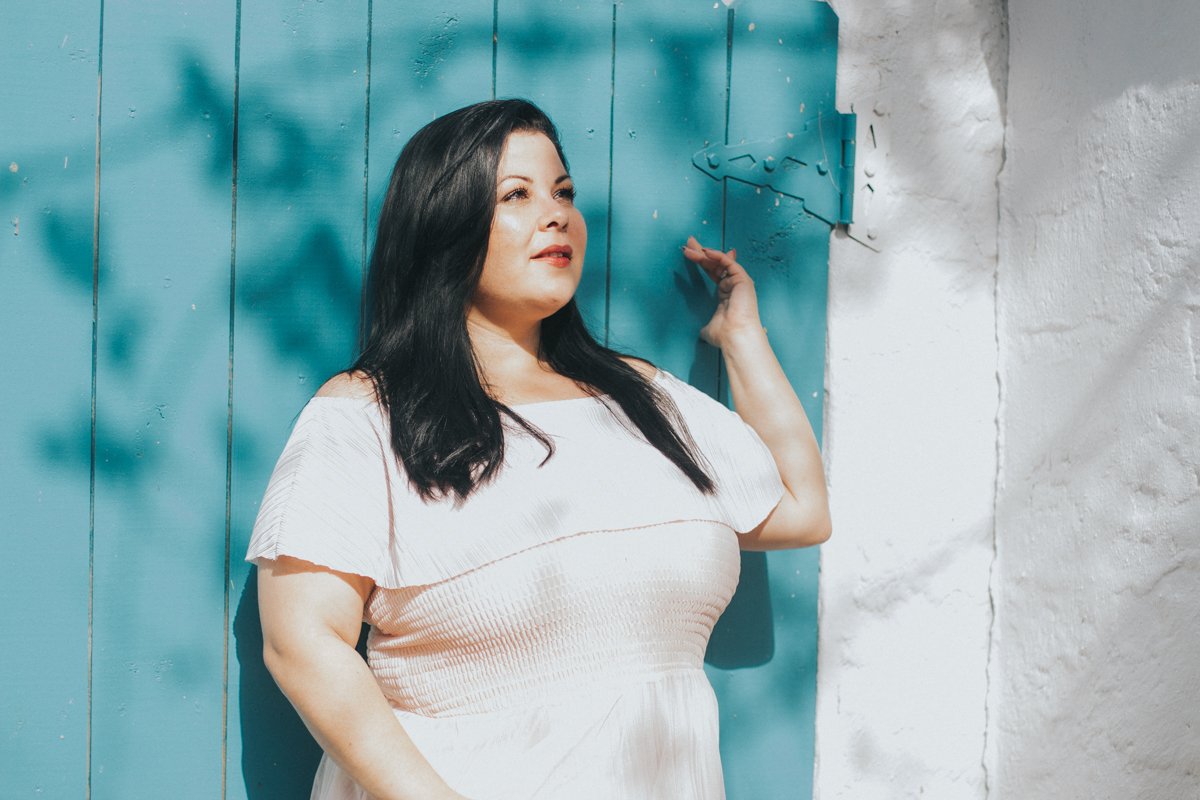 A plus-sized woman with dark hair wearing a white dress stands against a blue wooden wall, looking thoughtfully into the distance with her arm raised and resting on a hook.