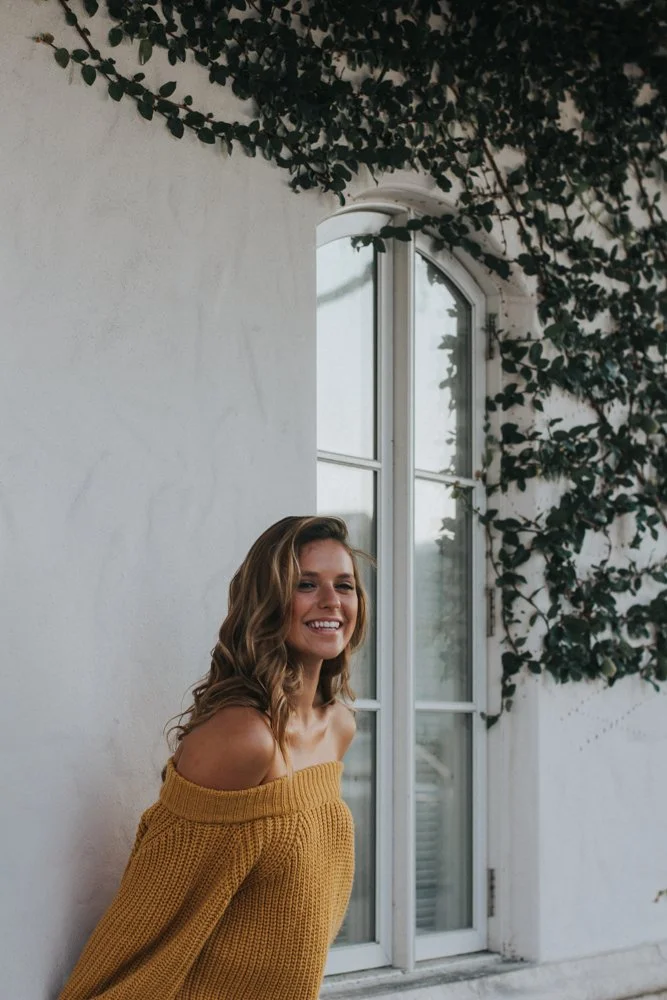 A woman with wavy blonde hair smiling and wearing a yellow off-the-shoulder sweater, standing in front of a white wall with a window and ivy growing on the wall.